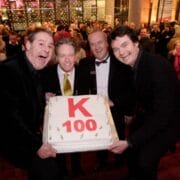 The panto cast hold a large cake with "K 100" written on it, standing on stairs in a crowded event venue decorated with lights.