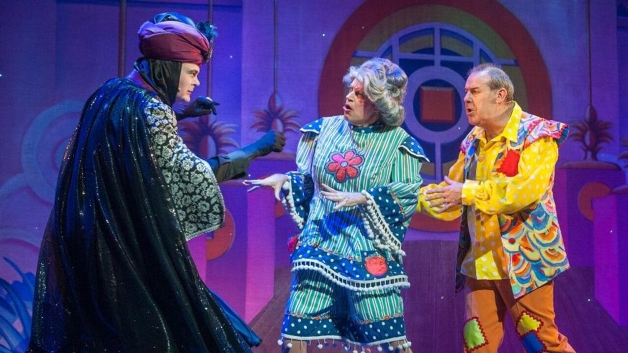 Three actors in colorful and elaborate costumes perform on stage, with a pink and blue whimsical set in the background.