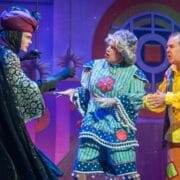 Three actors in colorful and elaborate costumes perform on stage, with a pink and blue whimsical set in the background.