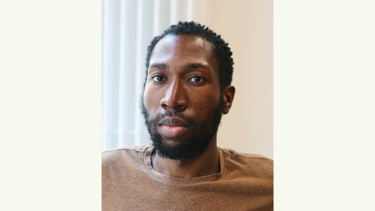 A man with short curly hair and a beard looks at the camera, wearing a brown shirt, with a neutral expression in a softly lit indoor setting.