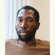 A man with short curly hair and a beard looks at the camera, wearing a brown shirt, with a neutral expression in a softly lit indoor setting.