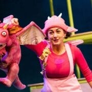 Bathed in the spotlight, a performer in a pink costume and hat holds a pink dragon puppet on stage—a whimsical scene reminiscent of Julia Donaldson’s beloved tales, with a set piece and another person visible in the background.