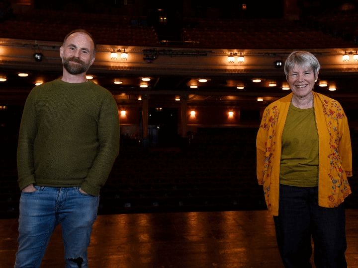A man (Laurie Sansom) and and a woman (Rhona Munro) stand on the Festival Theatrer stage facing the camera, with seats and theater lights visible in the background.