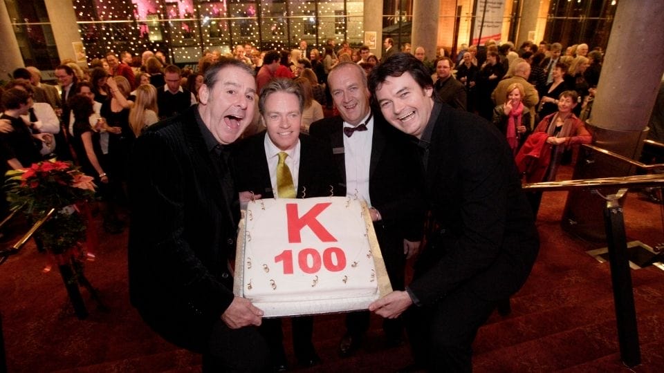 The panto cast hold a large cake with "K 100" written on it, standing on stairs in a crowded event venue decorated with lights.