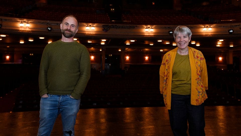 A man (Laurie Sansom) and and a woman (Rhona Munro) stand on the Festival Theatrer stage facing the camera, with seats and theater lights visible in the background.
