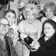 A group of five people pose indoors; one person in costume holds a champagne bottle, while others smile for the camera.