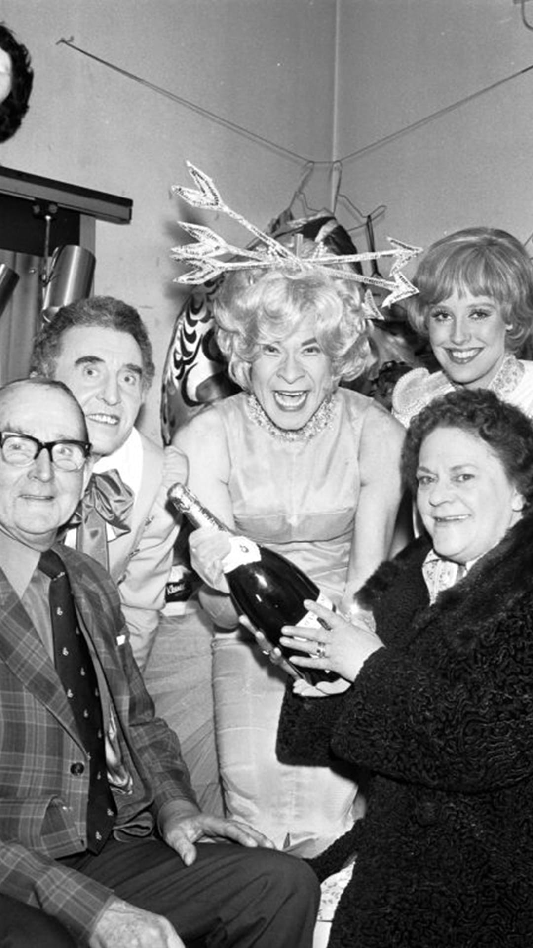 A group of five people pose indoors; one person in costume holds a champagne bottle, while others smile for the camera.