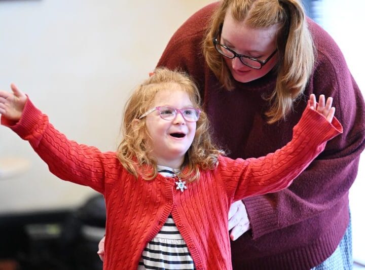 A young girl with glasses and a red sweater raises her arms while an adult woman beside her watches and smiles.