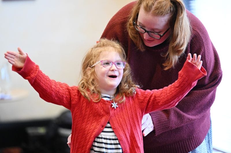 A young girl with glasses and a red sweater raises her arms while an adult woman beside her watches and smiles.