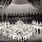 A large group of performers in uniform pose in formation on stage with curtains, promoting "The Gang Show 1960" at King's Theatre, as shown in the event poster inset.