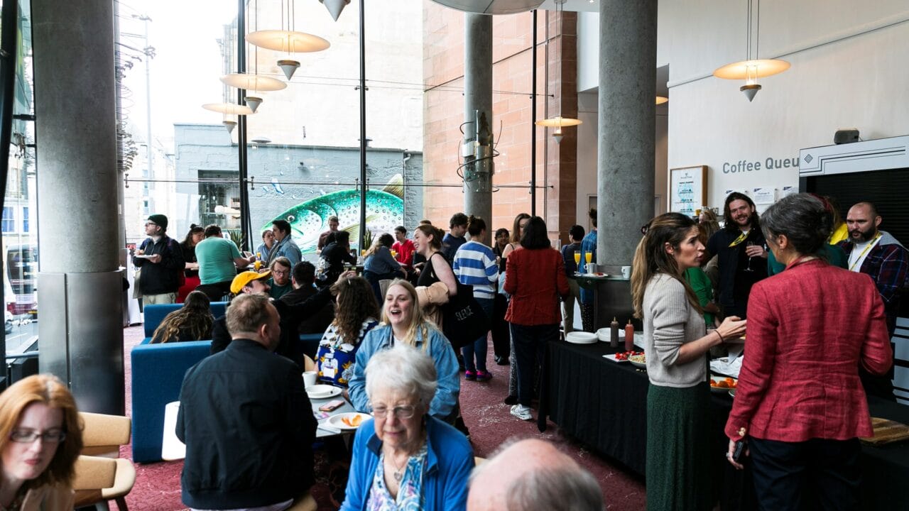 A group of people socializes and enjoys refreshments in a modern indoor space with large windows and a coffee queue sign.