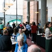 A group of people socializes and enjoys refreshments in a modern indoor space with large windows and a coffee queue sign.