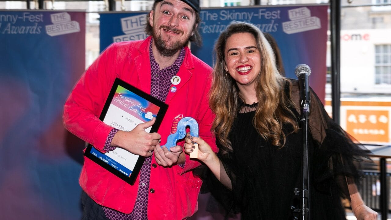 Two people stand indoors, smiling at the camera. One holds a framed certificate, the other holds a blue award ribbon. A microphone is visible in front of them.