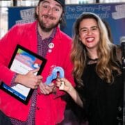 Two people stand indoors, smiling at the camera. One holds a framed certificate, the other holds a blue award ribbon. A microphone is visible in front of them.