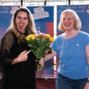 Two women smiling; one in black holding a bouquet of yellow and pink flowers, the other in blue. Event banners for "The Skinny-Fest Festival Awards" are visible in the background.