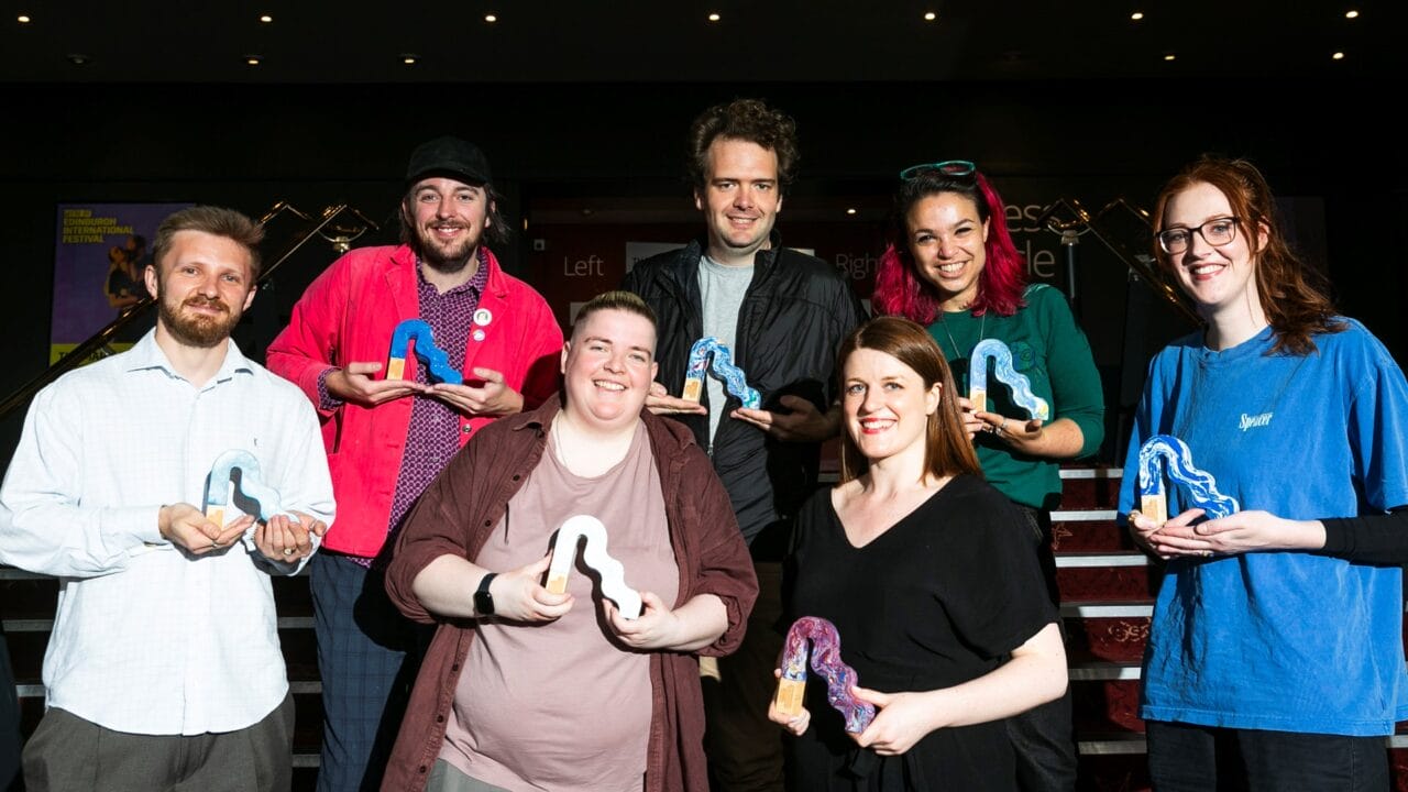 Seven people stand in two rows, each holding a colorful abstract-shaped award, posing and smiling for a group photo indoors under bright lighting.