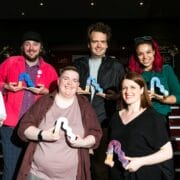 Seven people stand in two rows, each holding a colorful abstract-shaped award, posing and smiling for a group photo indoors under bright lighting.