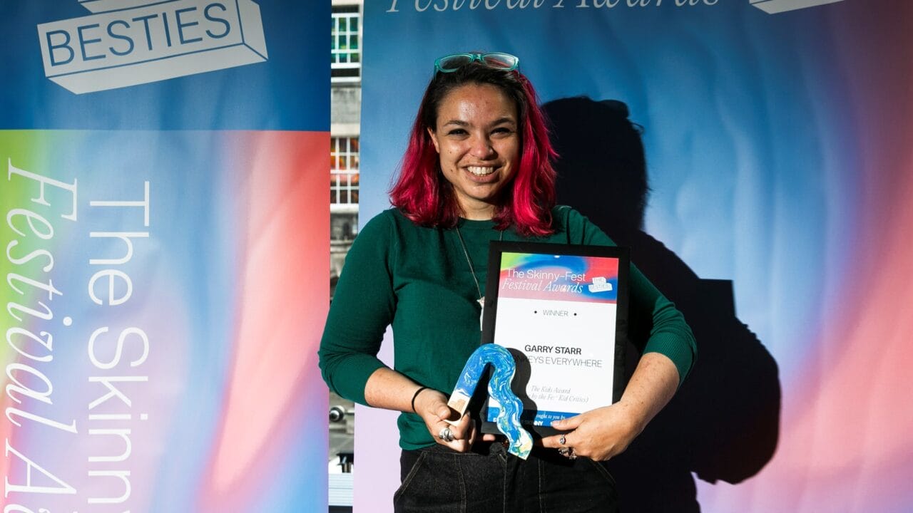 A person with pink hair, wearing glasses and a green top, holds an award and certificate in front of a colorful "Festival Awards" banner.