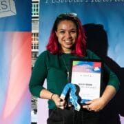 A person with pink hair, wearing glasses and a green top, holds an award and certificate in front of a colorful "Festival Awards" banner.