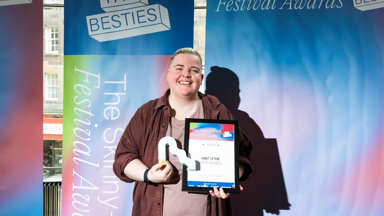 Person smiling and holding an award trophy and a framed certificate in front of a colorful "Festival Awards" backdrop.