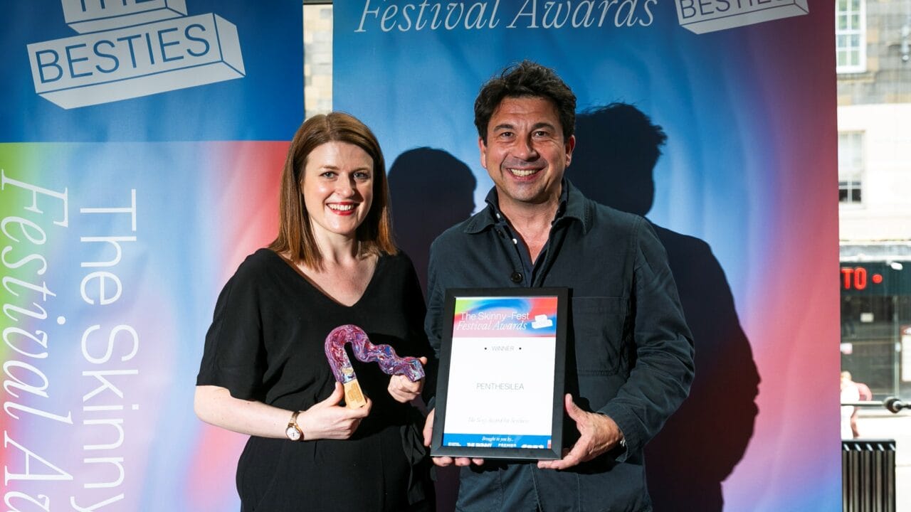 Two people stand indoors, smiling and holding a colorful trophy and an award plaque in front of a "Festival Awards" banner.
