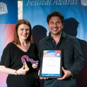 Two people stand indoors, smiling and holding a colorful trophy and an award plaque in front of a "Festival Awards" banner.