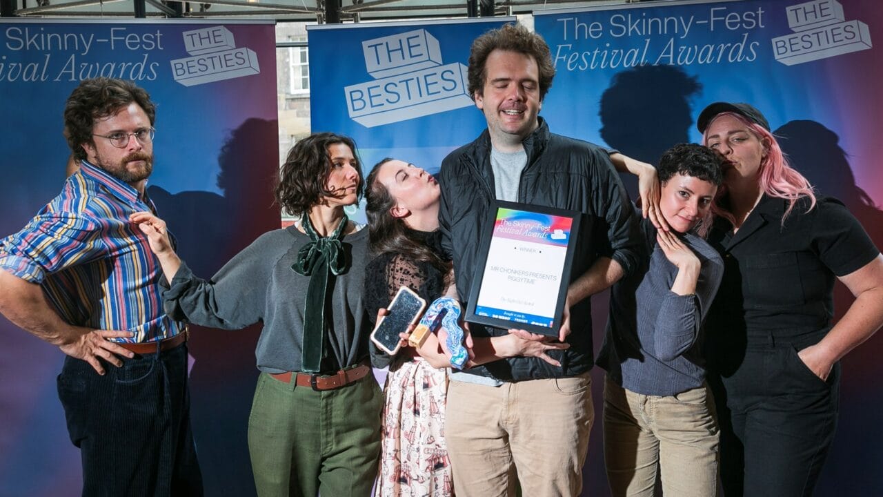 Six people pose together at The Skinny-Fest Festival Awards, with one holding a framed award and others displaying celebratory expressions in front of event banners.