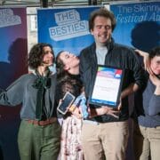 Six people pose together at The Skinny-Fest Festival Awards, with one holding a framed award and others displaying celebratory expressions in front of event banners.