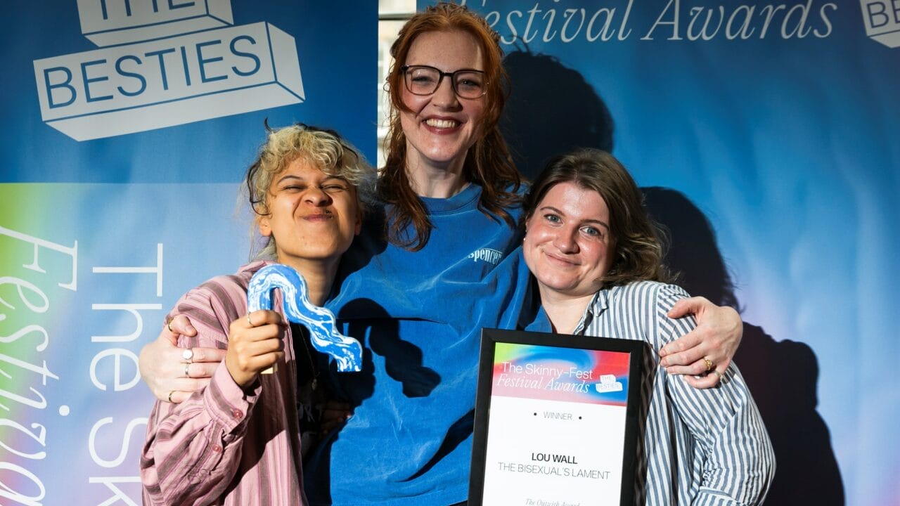Three people stand together smiling; one holds a wavy striped award, another holds a framed certificate labeled "Winner." Blue festival banners are visible in the background.