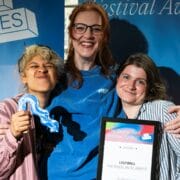 Three people stand together smiling; one holds a wavy striped award, another holds a framed certificate labeled "Winner." Blue festival banners are visible in the background.