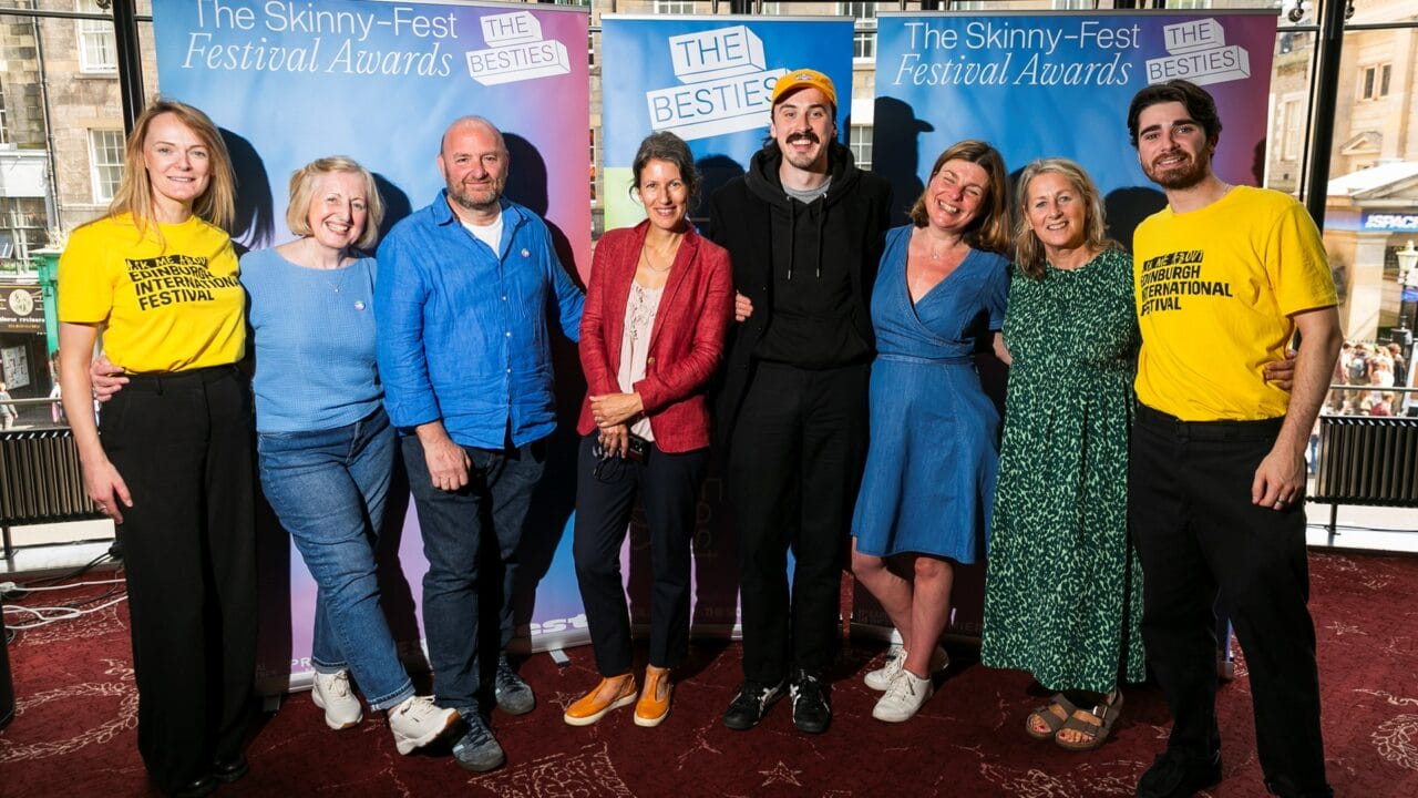 Eight people stand together indoors in front of two Skinny-Fest Festival Awards banners, posing for a group photo.