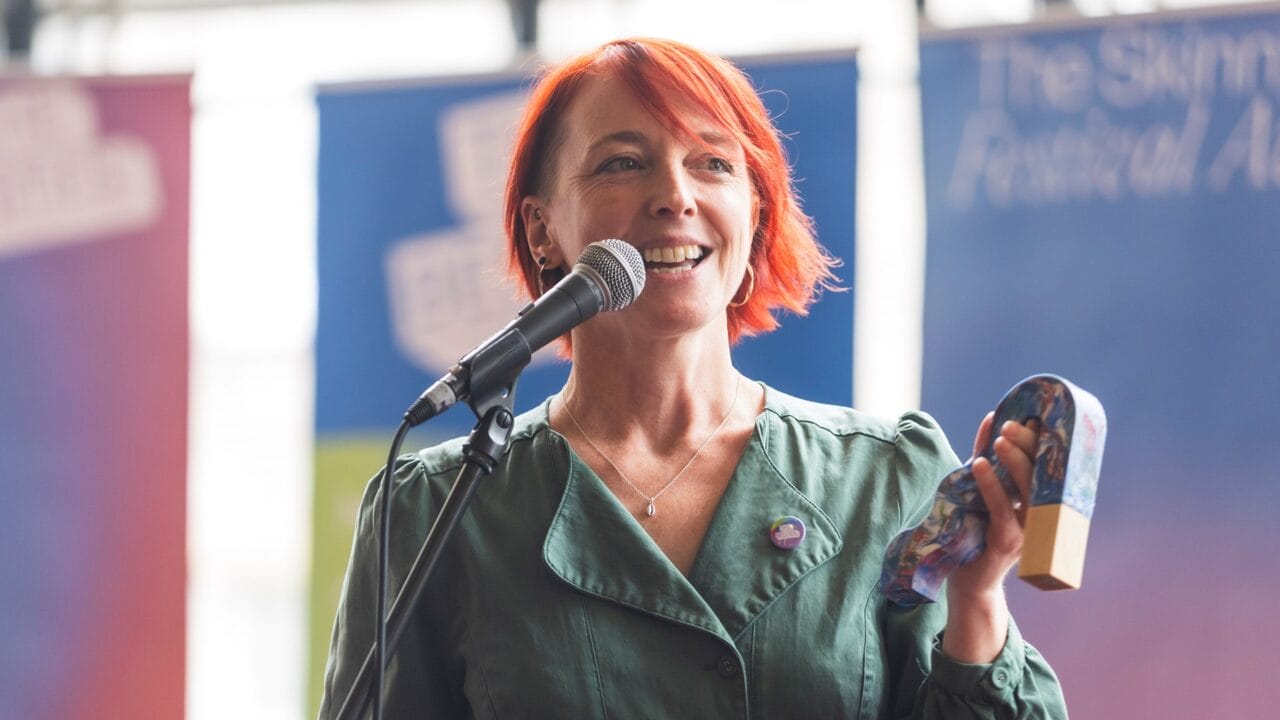 A woman with short red hair speaks into a microphone while holding an award, standing in front of colorful banners.