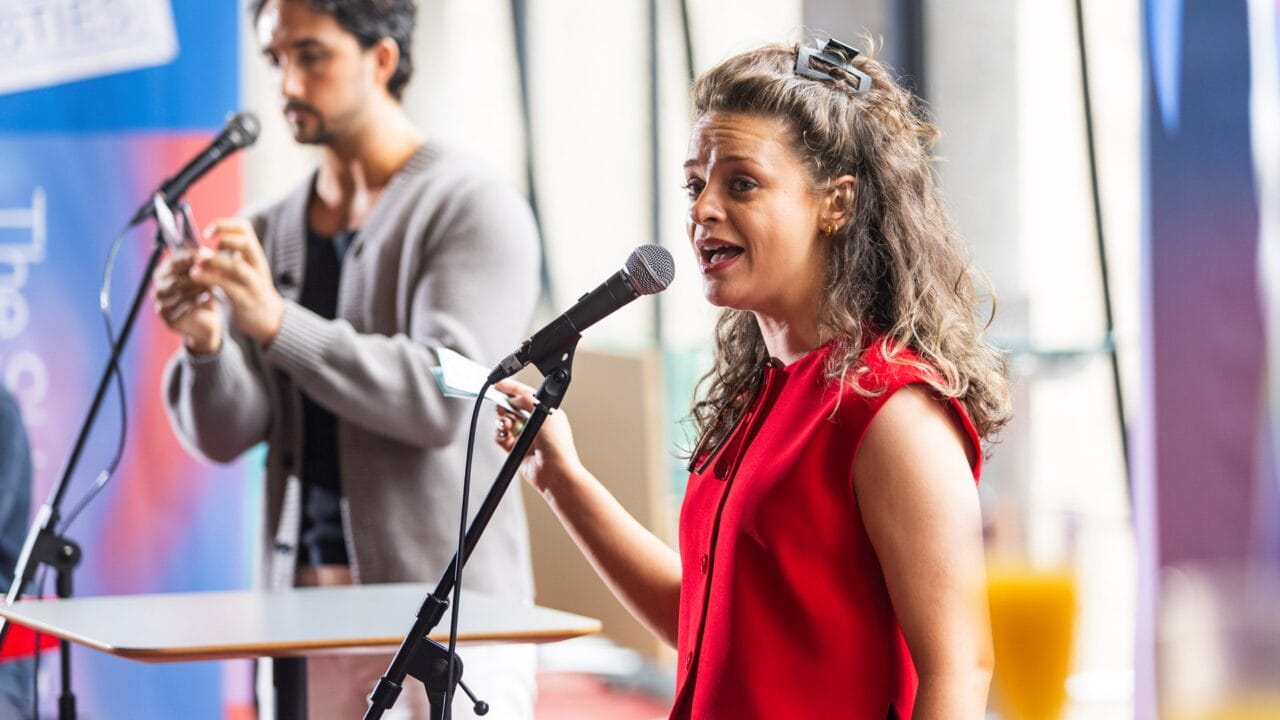 A woman in a red sleeveless top speaks or sings into a microphone, while a man behind her appears to use a smartphone near another microphone.