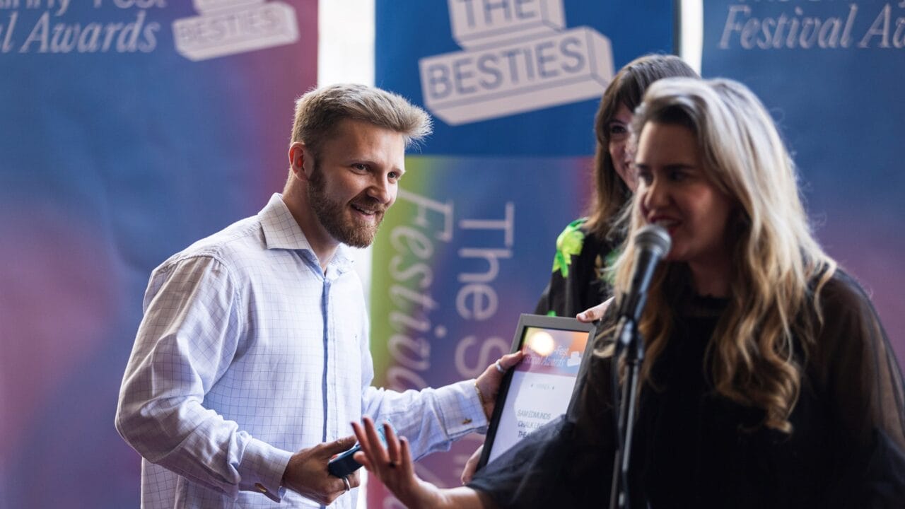 A man in a white shirt smiles and accepts a framed certificate from a woman speaking into a microphone at an awards event.