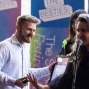 A man in a white shirt smiles and accepts a framed certificate from a woman speaking into a microphone at an awards event.