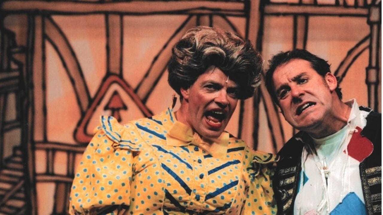 Two actors, Allan Stewart and Andy Gray, in colorful costumes perform on stage, Allan is in Dame costume wearing a yellow polka-dot dress and wig, both have animated facial expressions against a painted backdrop.