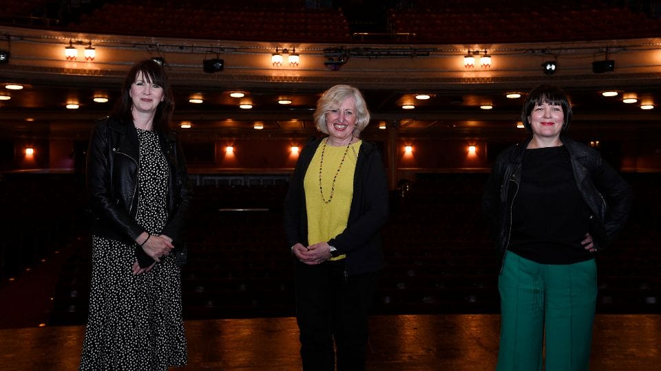 Three women stand on a theater stage, with Fiona Gibson in the centre, with auditorium seats and warm lighting in the background.