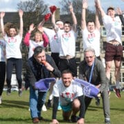 A group of Capital Theatres staff wearing matching t-shirts jump in the air, while three men in front pose with foam hands on the grass at Bruntsfield Links.