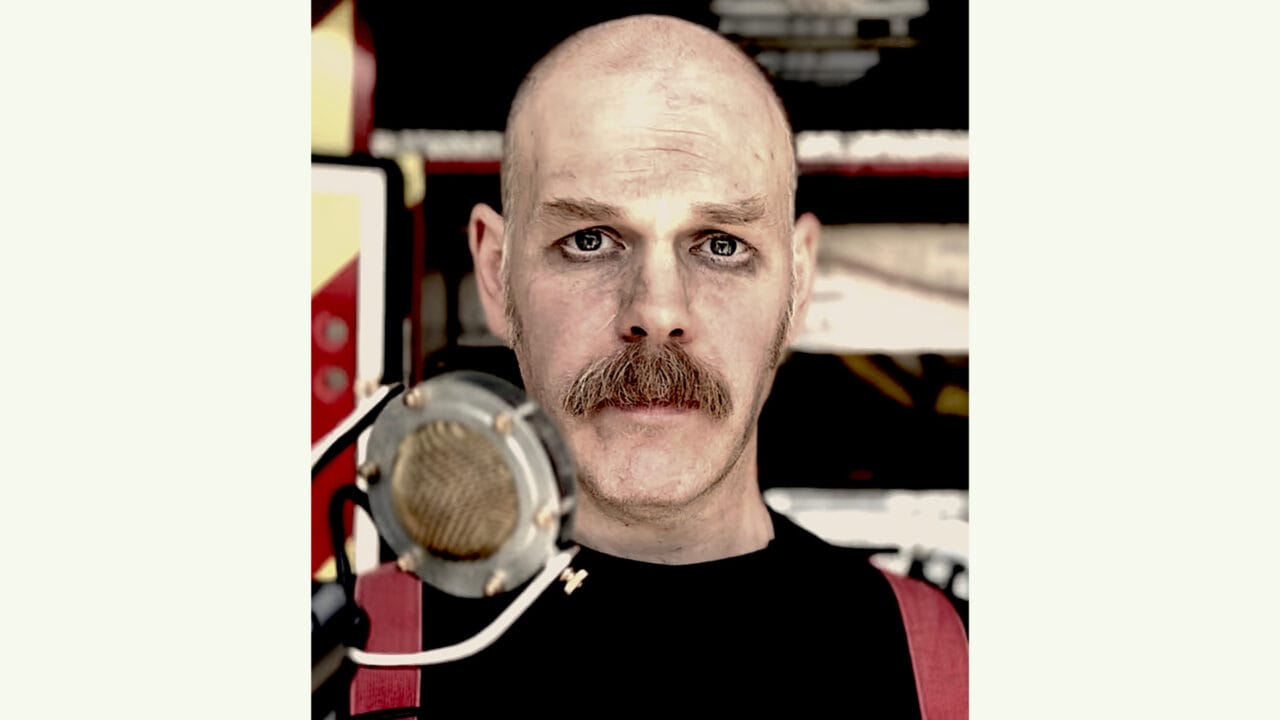 A bald man with a moustache and dark eye makeup stands in front of a vintage microphone, wearing a black shirt and red suspenders.