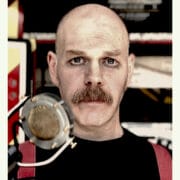A bald man with a moustache and dark eye makeup stands in front of a vintage microphone, wearing a black shirt and red suspenders.