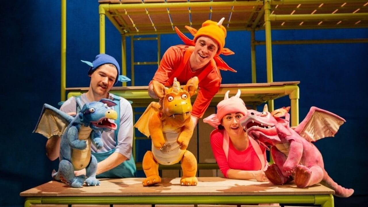 Three actors in colorful costumes operate blue, orange, and pink dragon puppets under the spotlight on a stage set with green scaffolding, bringing a Julia Donaldson story to life.