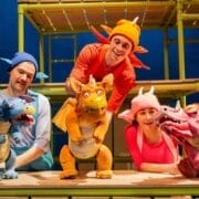 Three actors in colorful costumes operate blue, orange, and pink dragon puppets under the spotlight on a stage set with green scaffolding, bringing a Julia Donaldson story to life.