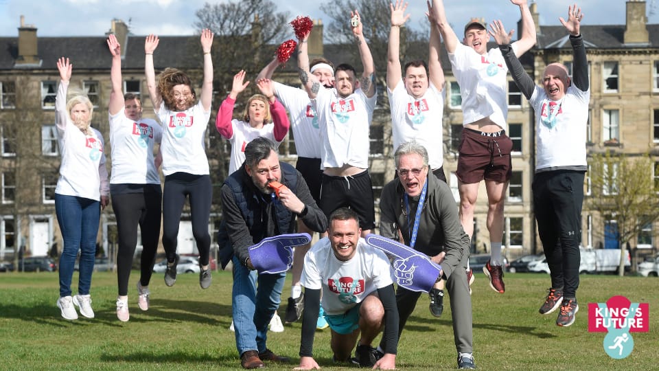 A group of Capital Theatres staff wearing matching t-shirts jump in the air, while three men in front pose with foam hands on the grass at Bruntsfield Links.