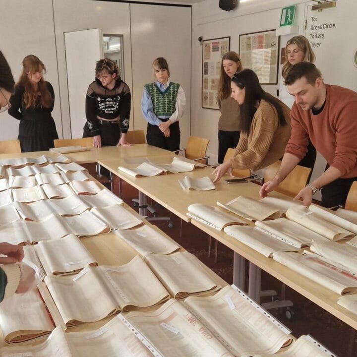 Eight people stand around a table examining and organizing rows of open archival books or documents in a well-lit room.