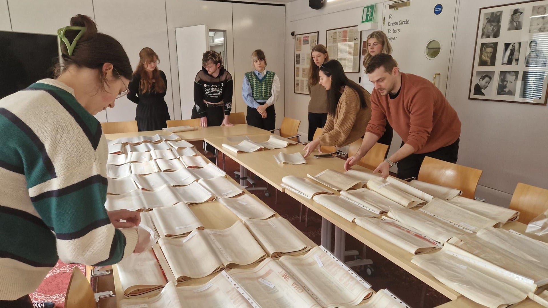 Eight people stand around a table examining and organizing rows of open archival books or documents in a well-lit room.