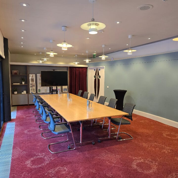 A modern conference room with a long wooden table, office chairs, water pitchers, a TV screen, and bookshelves, set on red carpet with overhead lighting.