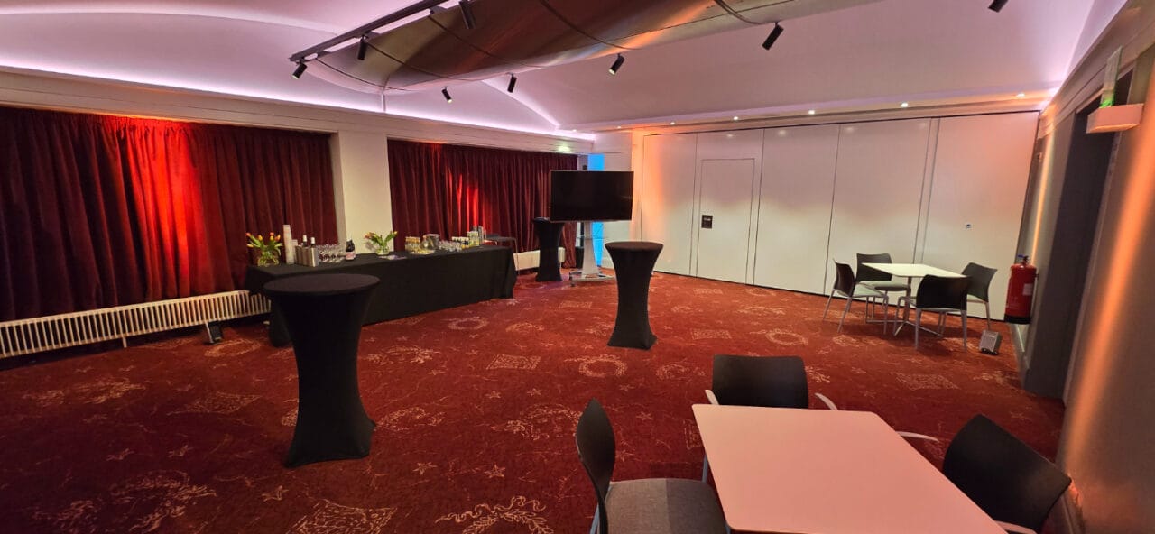 An event room with red carpet, round high tables, square dining tables with chairs, a buffet table with drinks and snacks, and red curtains along one wall.