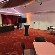 An event room with red carpet, round high tables, square dining tables with chairs, a buffet table with drinks and snacks, and red curtains along one wall.