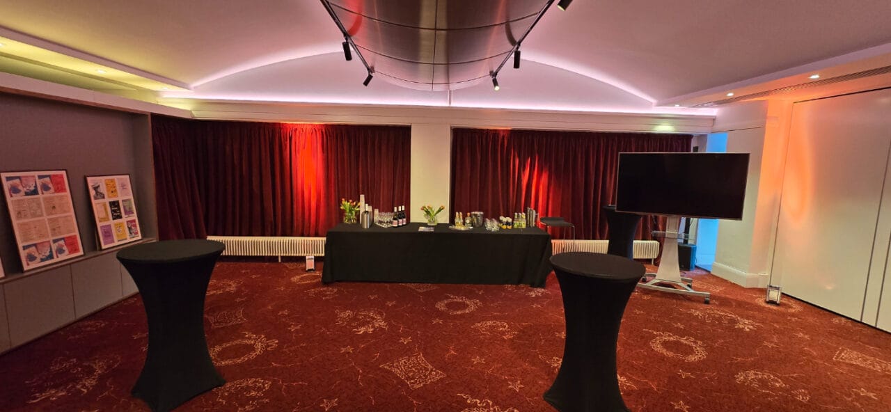 An event room with red carpet and curtains, a black-clothed table set with drinks, two cocktail tables, flower vases, framed posters, and a large TV screen.
