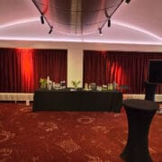 An event room with red carpet and curtains, a black-clothed table set with drinks, two cocktail tables, flower vases, framed posters, and a large TV screen.